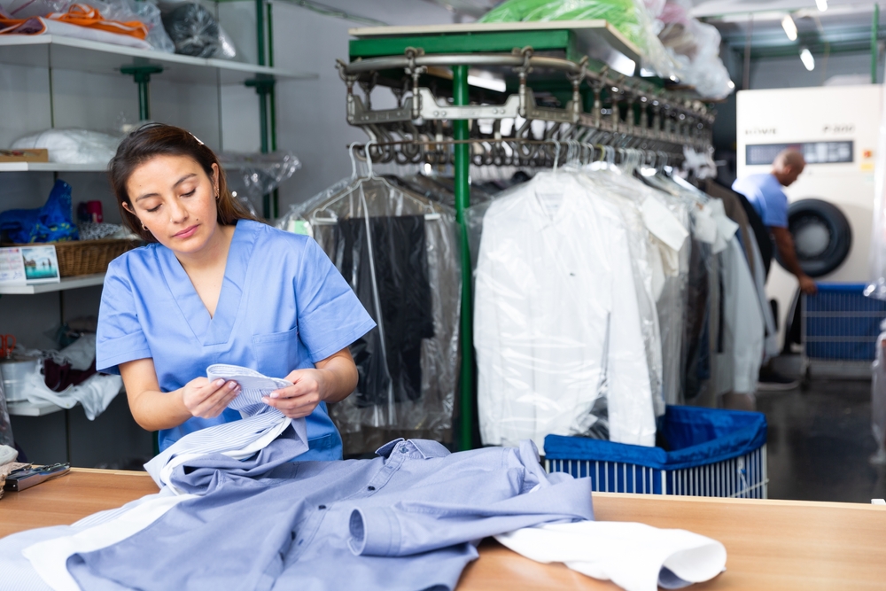 A woman in scrubs folds clean shirts at a dry cleaning facility, with hanging garments and laundry equipment visible in the background.
